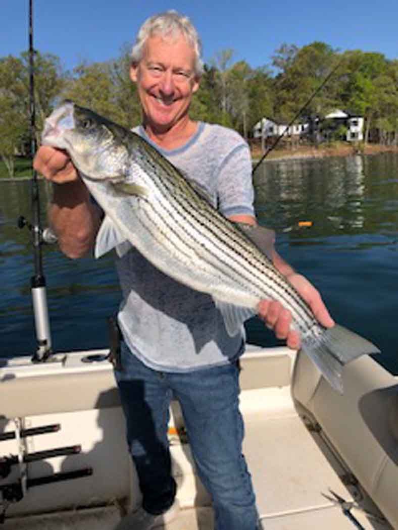 Man smiling on a boat holding a large striped fish by the lake