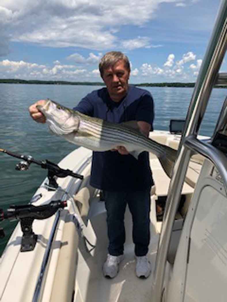 Man holding a large striped fish on a boat deck with calm water and sky in the background