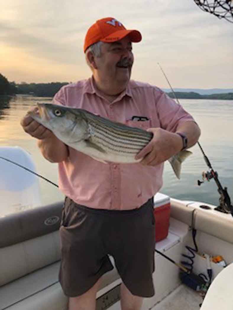 Man holding a large striped fish on a boat at sunset, wearing an orange cap and pink shirt