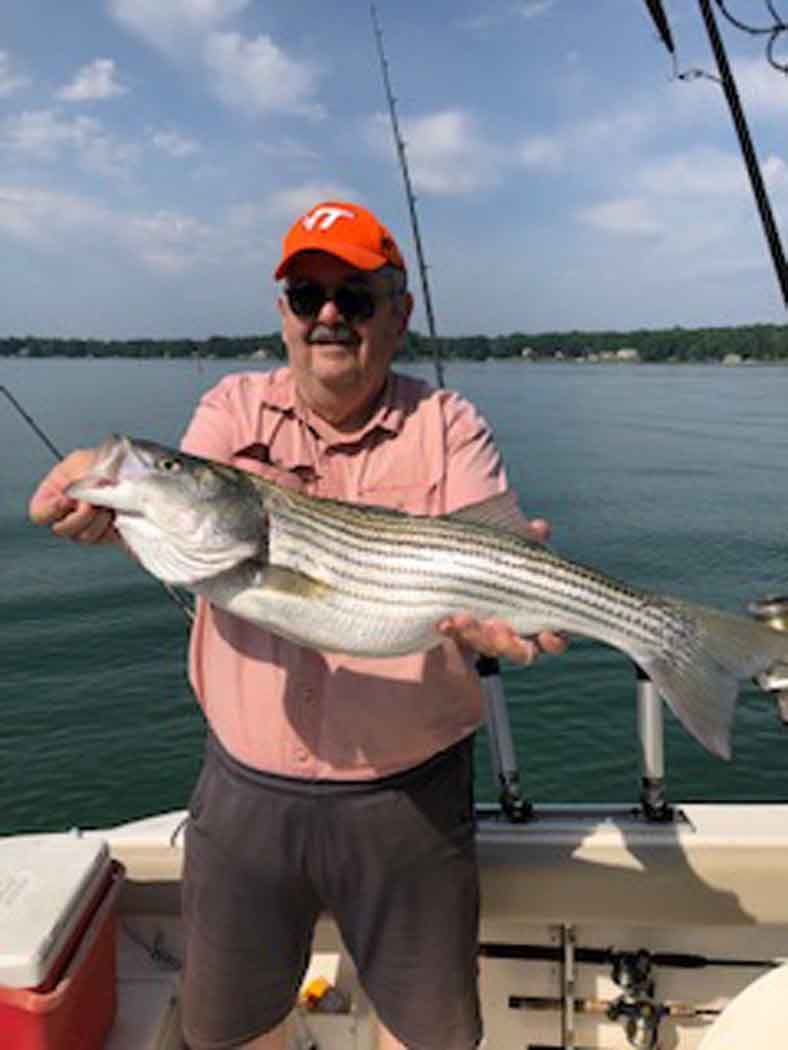 Man holding a large striped fish on a boat, with calm water and shoreline in the background.