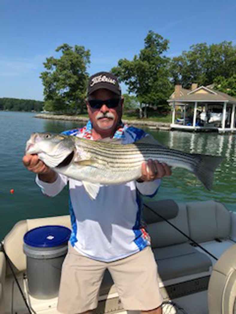 Man holding a large striped fish on a boat by a lake, with trees and a dock in the background.