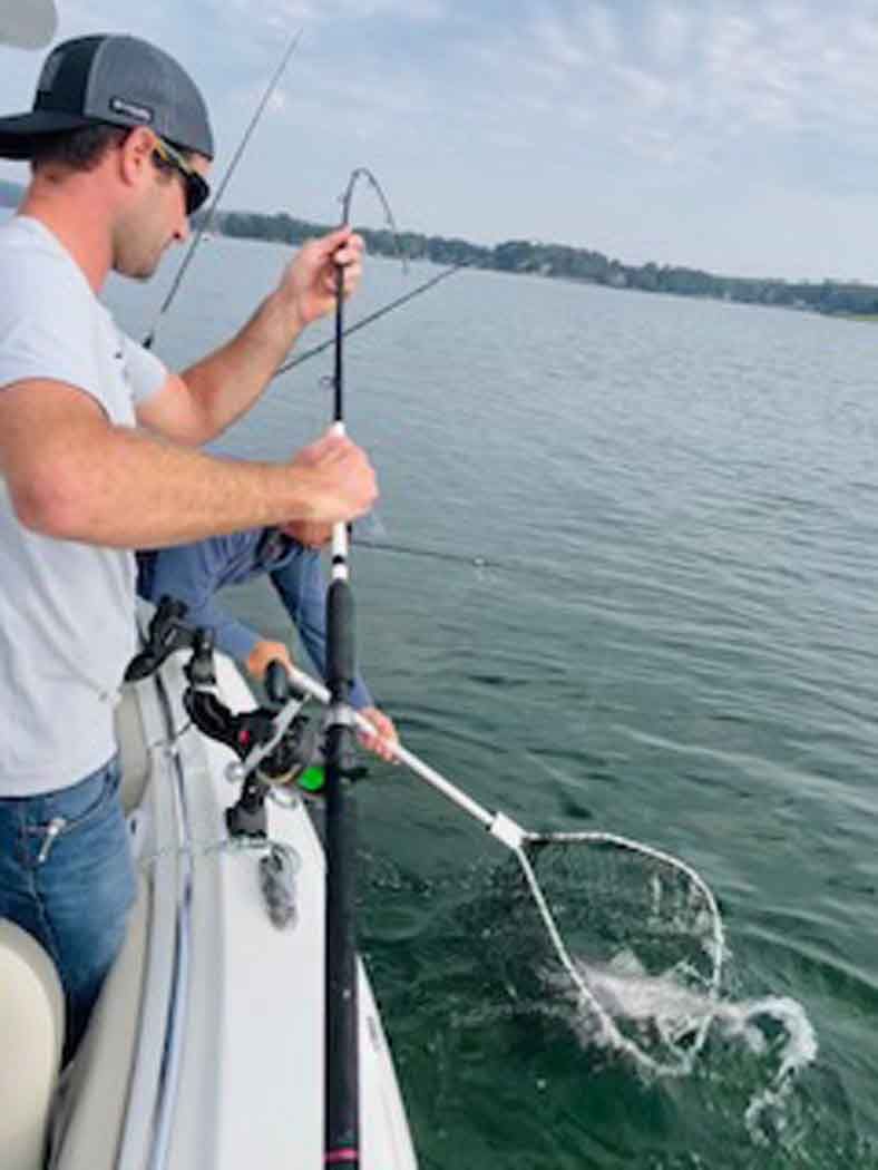 Person fishing from a boat, pulling a hooked fish from the water with a net.
