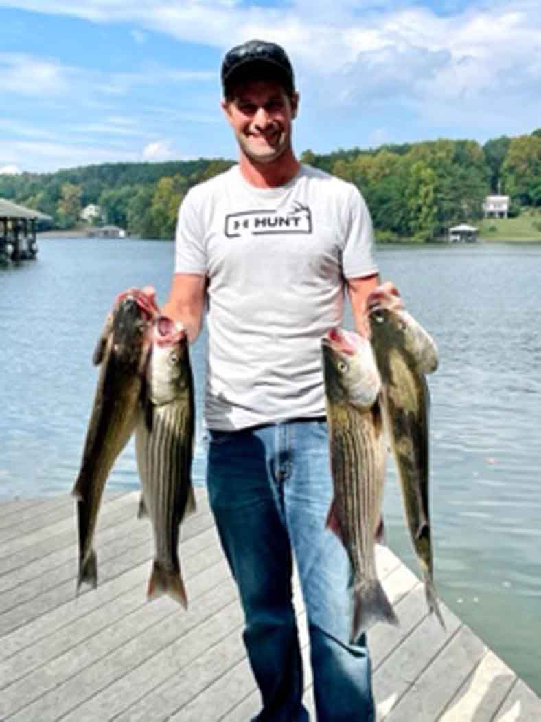 Man standing on a dock holding three large fish by a lake on a sunny day