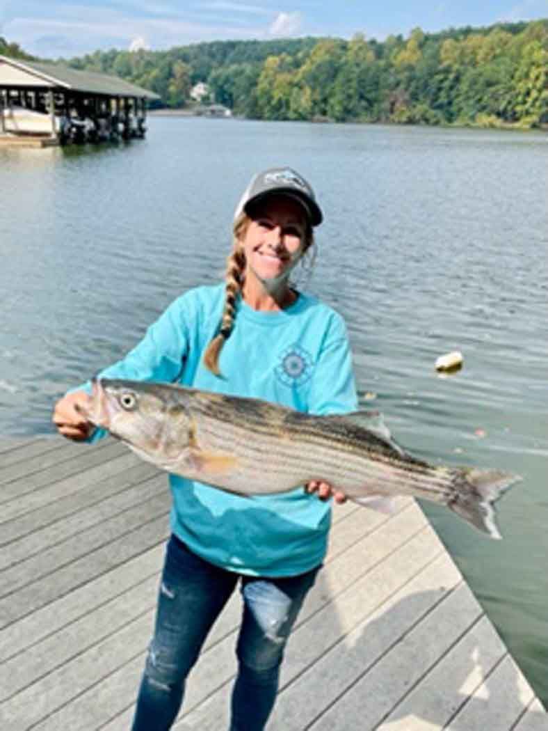 Woman in turquoise shirt holding a large striped fish on a lakeside dock.