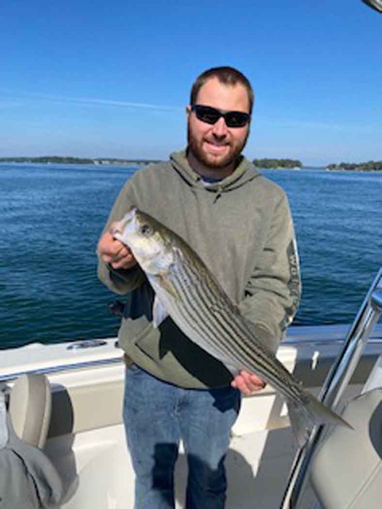 Man holding a striped fish on a boat with calm blue water and shoreline in the background