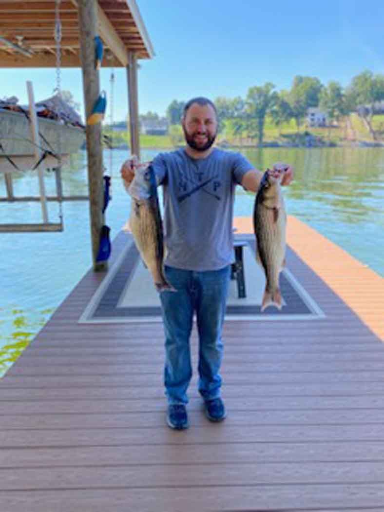 Man standing on a dock holding two large fish, with a lake and boats in the background.