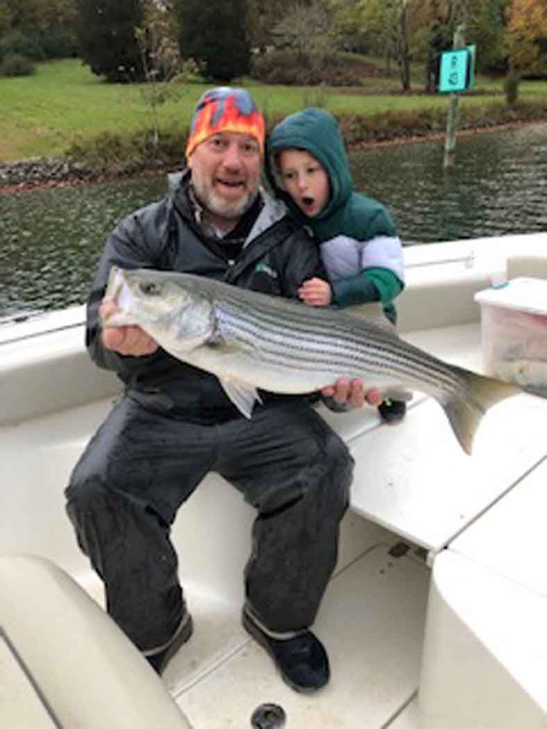 Man and child on a boat holding a large fish, with water and shoreline in the background.