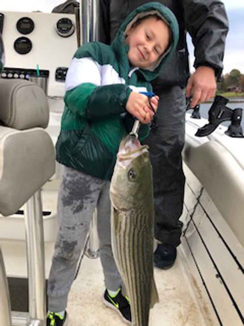 Smiling child holding a large fish on a boat deck, with an adult standing behind.