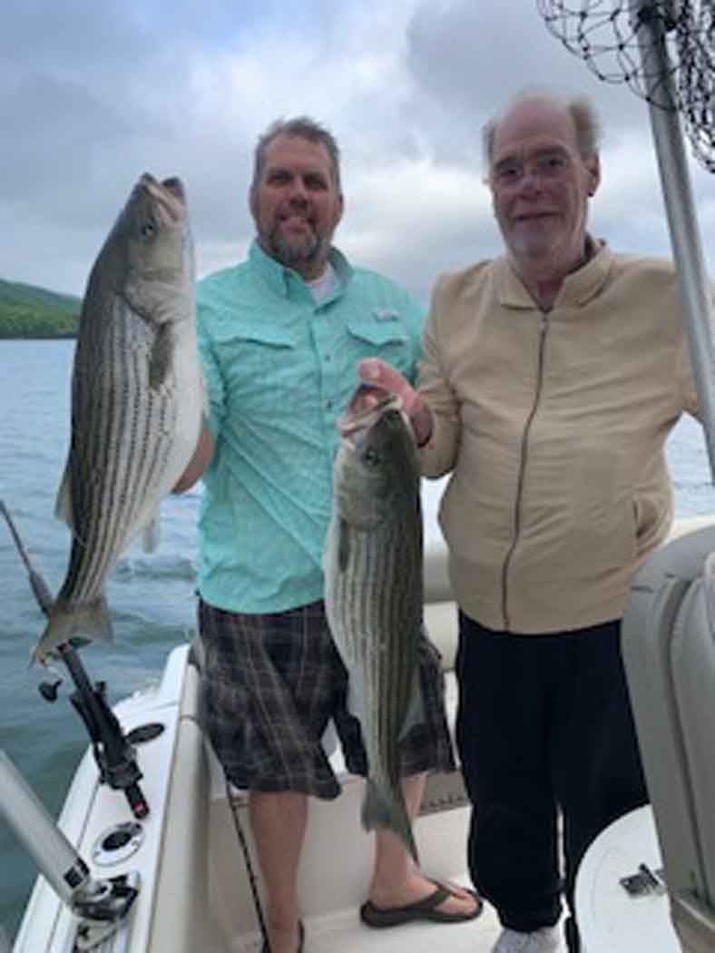 Two men on a boat holding striped fish, with water and hills in the background.