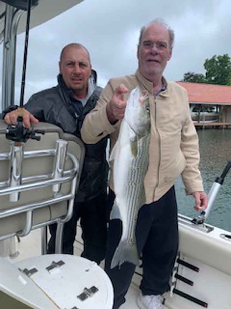 Two people on a boat hold a large striped fish near the water