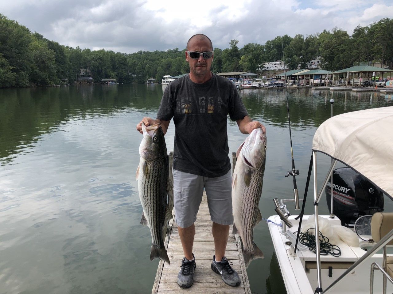 Man holding two large fish on a dock beside a calm lake with boats and trees in the background