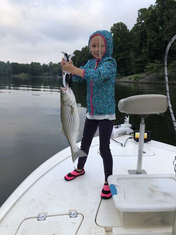 Child holding a large striped fish on a boat deck by a calm lake
