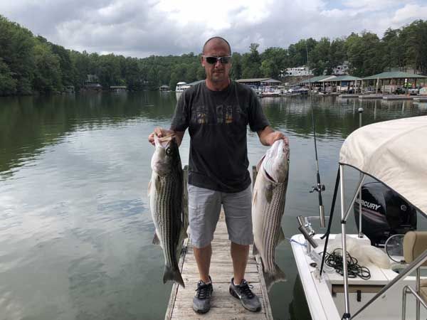 Man standing on a dock, holding two large fish beside a lake and boats under cloudy skies