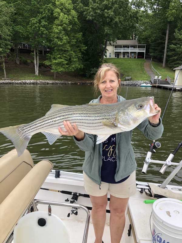 Woman holding a large striped fish on a boat docked by a calm lake with trees and a house in the background
