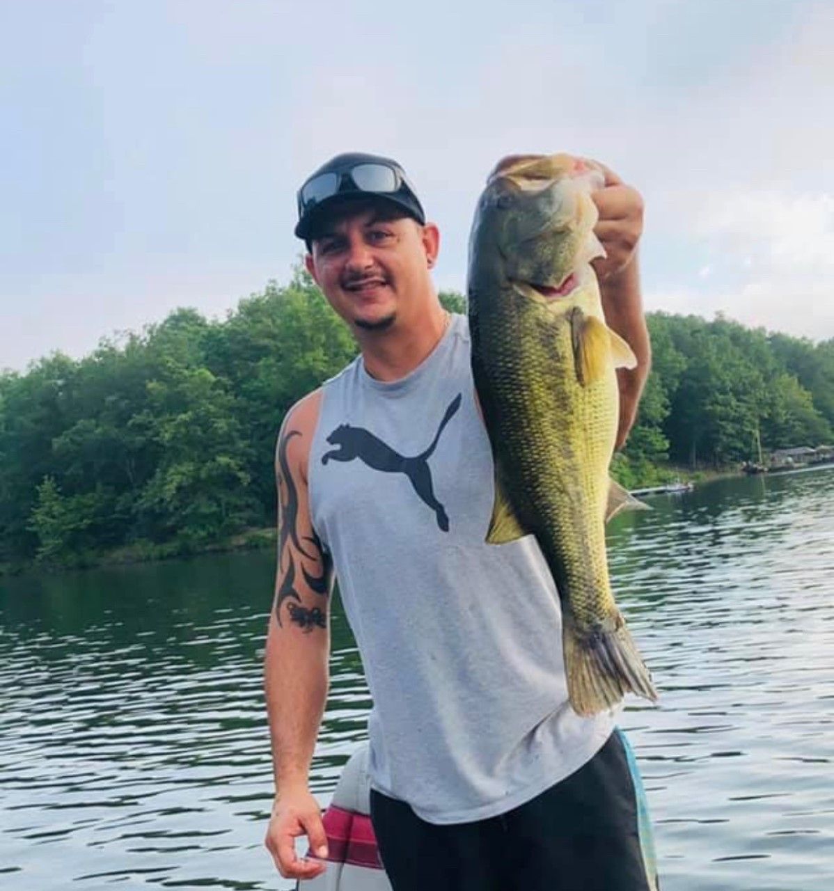 Man holding a large bass by a lake, smiling in a gray Puma shirt and black cap