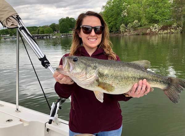 Woman in sunglasses holding a large fish on a boat with a calm lake and trees behind her