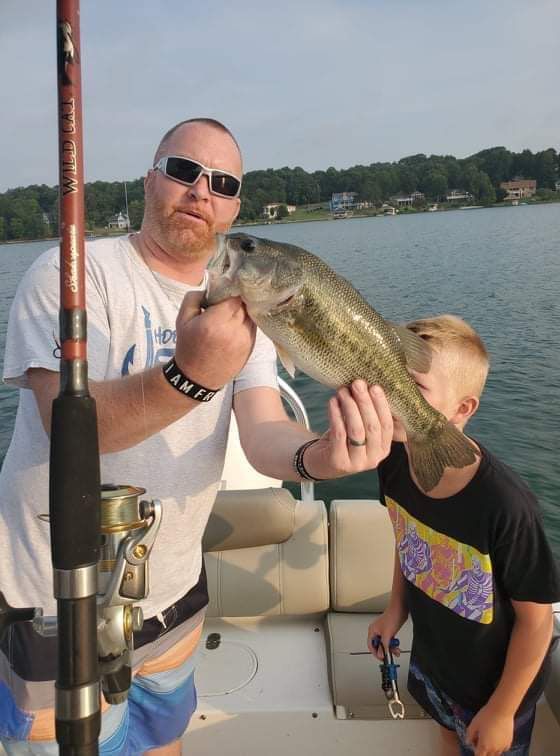 Two people on a boat hold up a large fish beside a lake at sunset.