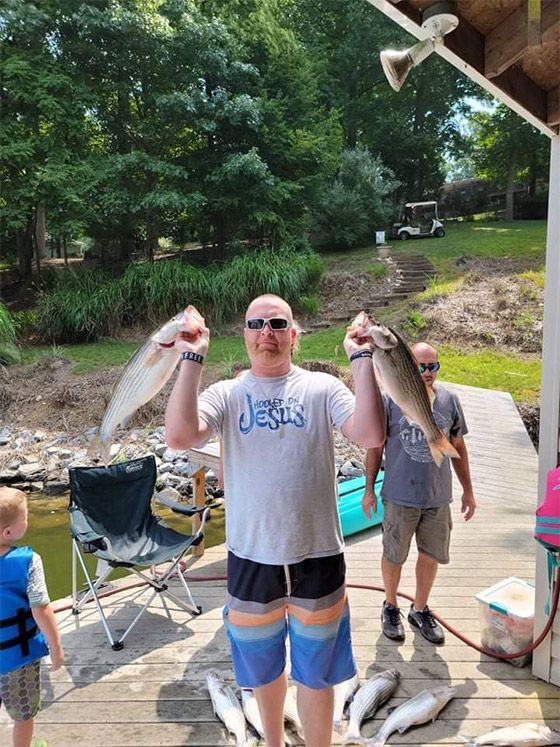 Two people holding large fish on a porch by a river, with a child nearby.