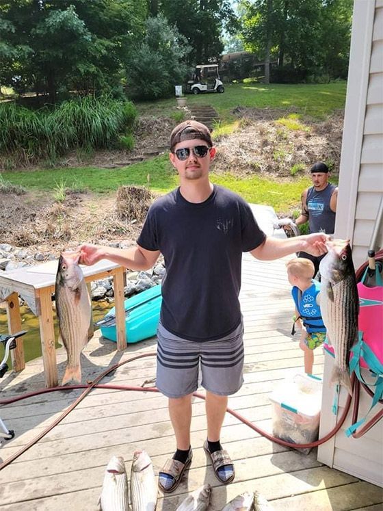 Person in sunglasses holding two large fish on a dock, with children and a river in the background