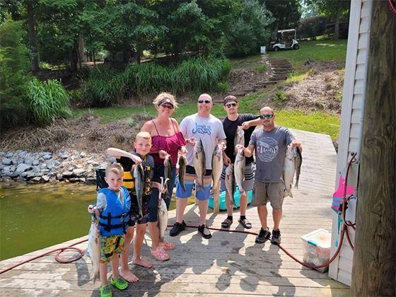Group of people posing with fishing gear on a dock beside a pond and wooded shoreline