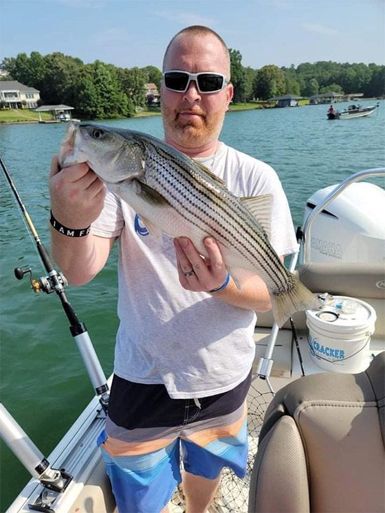 Man holding a striped fish on a boat on a lake, wearing sunglasses and a white shirt.