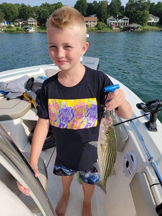 Boy on a boat holding a striped fish on a lake, with houses and trees in the background.