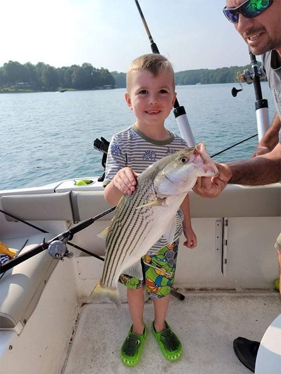 Child holding a large striped fish on a boat, with an adult nearby and a lake in the background