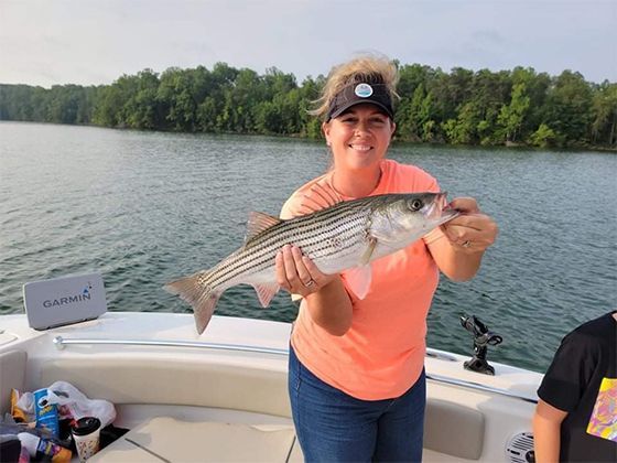Person on a boat holding a striped fish on a lake with trees in the background