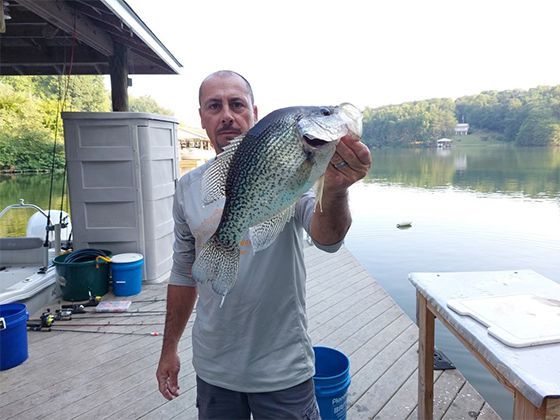 Man holding a large fish on a lakeside dock with buckets and a table nearby