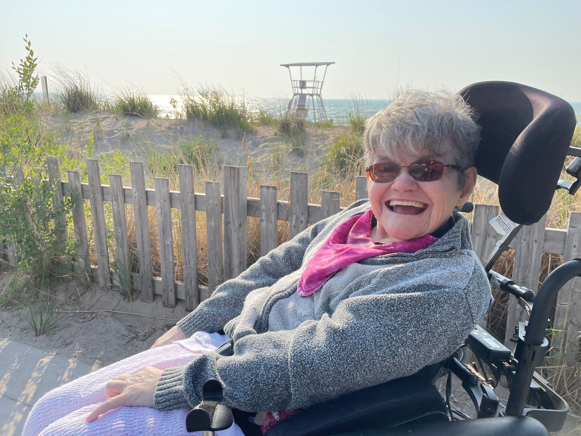 An elderly woman in a wheelchair is smiling in front of a lifeguard tower.
