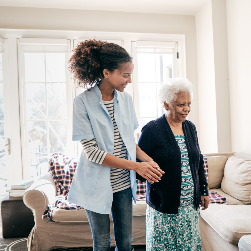 A young woman is helping an older woman walk in a living room.