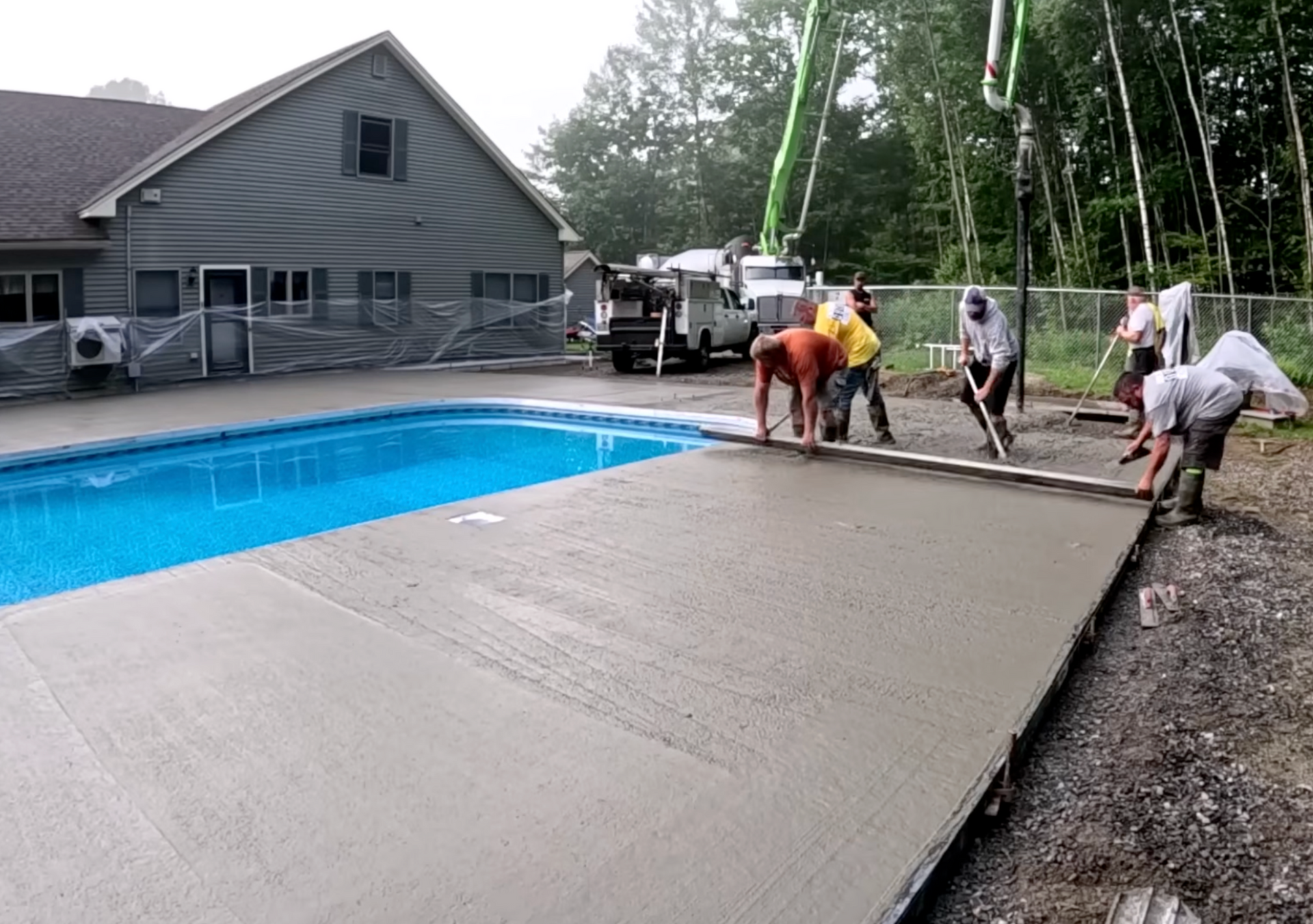 Concrete patio installation beside a residential swimming pool, with workers pouring and leveling fresh concrete slab for backyard outdoor space.