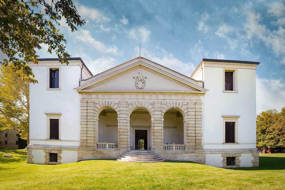 Edificio neoclassico bianco con colonne e due ali, situato su un prato erboso sotto un cielo azzurro.