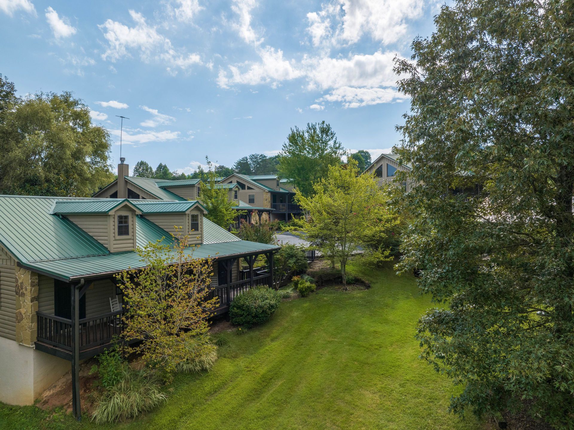 Green-roofed cabins on a grassy hillside, surrounded by trees under a partly cloudy sky.