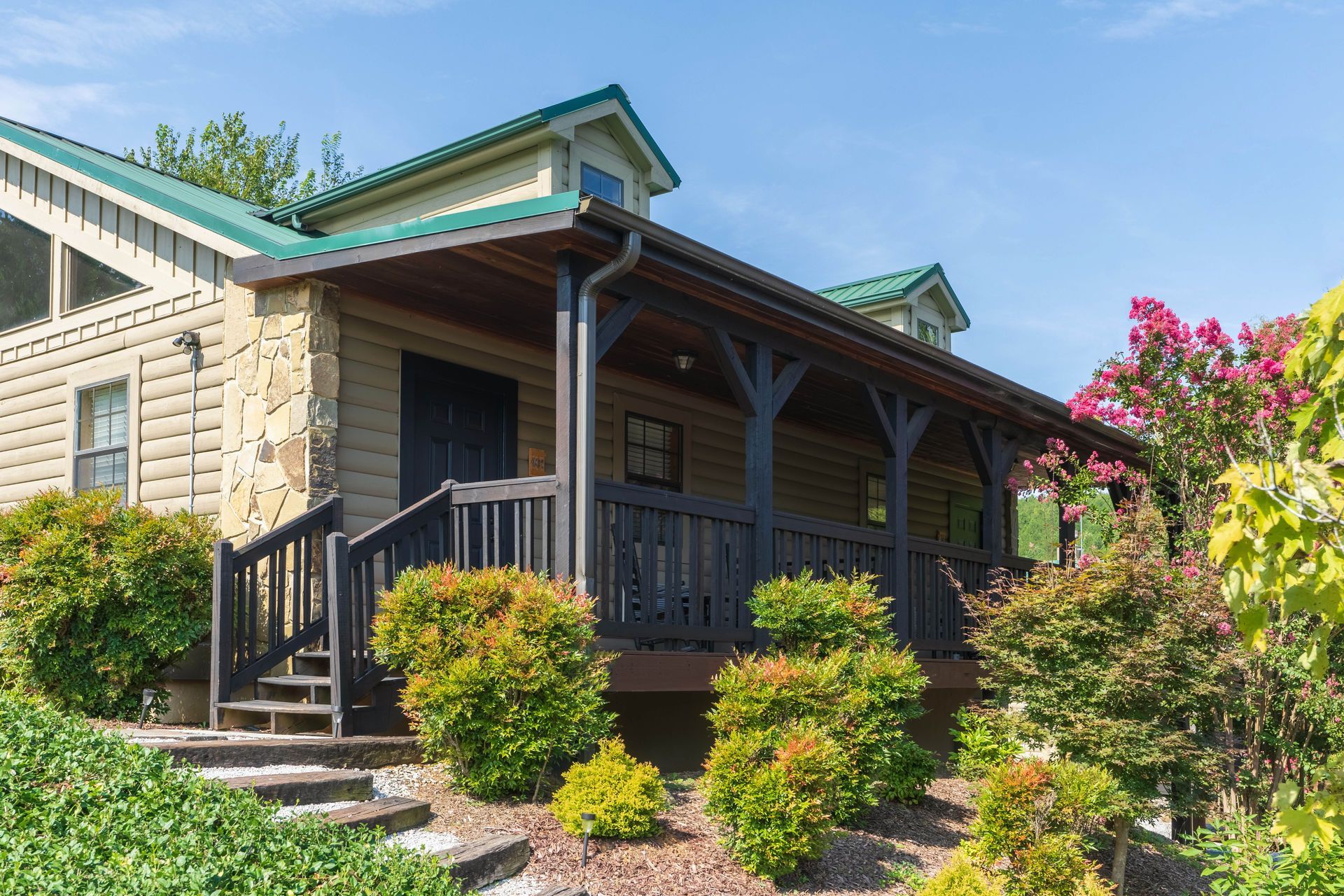Cabin with green roof and wooden porch, surrounded by greenery under a blue sky.