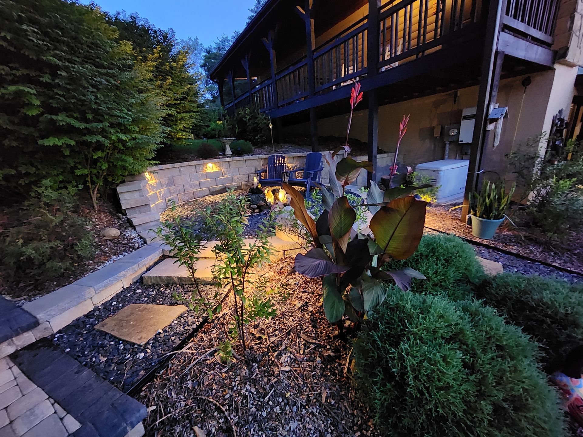 Lit backyard garden with retaining walls, a deck, plants, and a small water feature, at dusk.