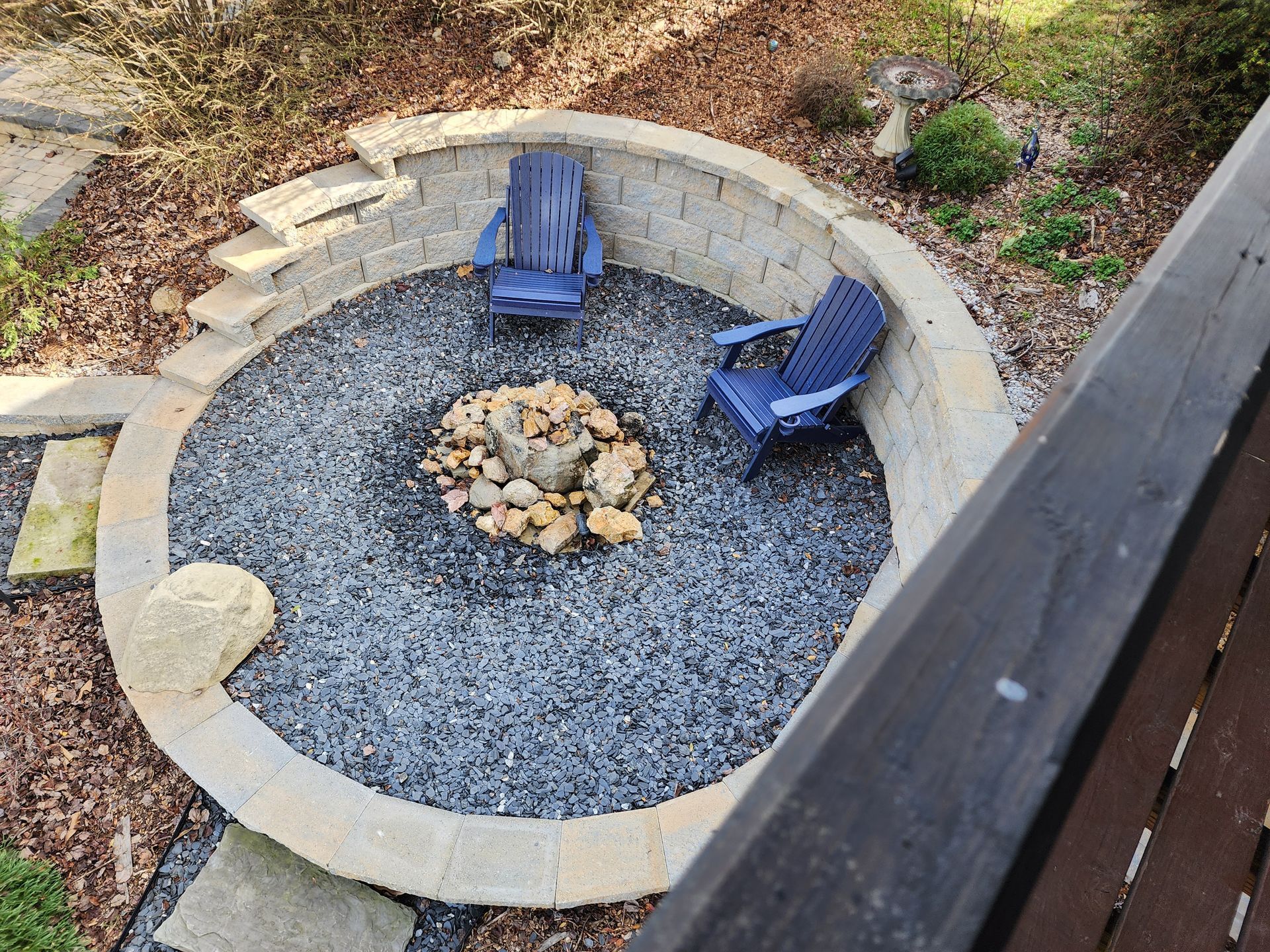 Fire pit area with two blue chairs, gravel ground, and stone wall in a yard.