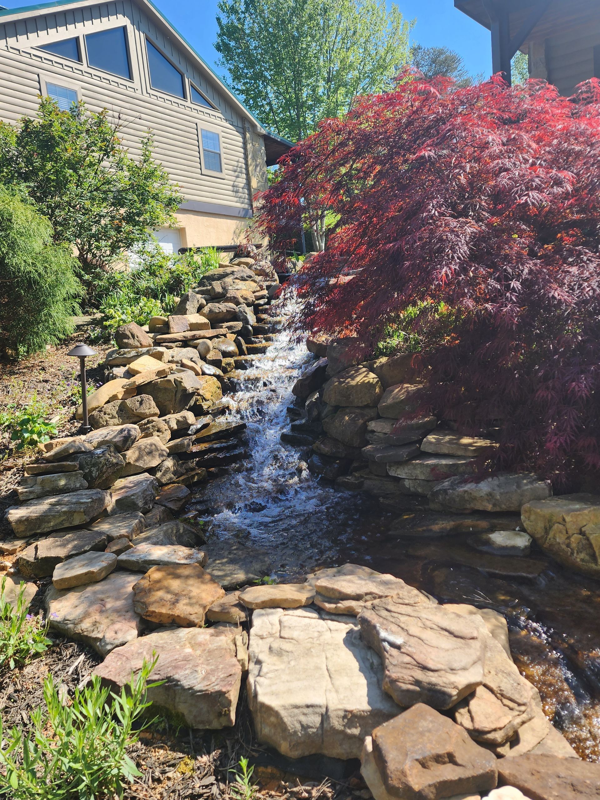Waterfall cascading down rocks, flanked by green and red foliage, next to a house with a sloped roof.