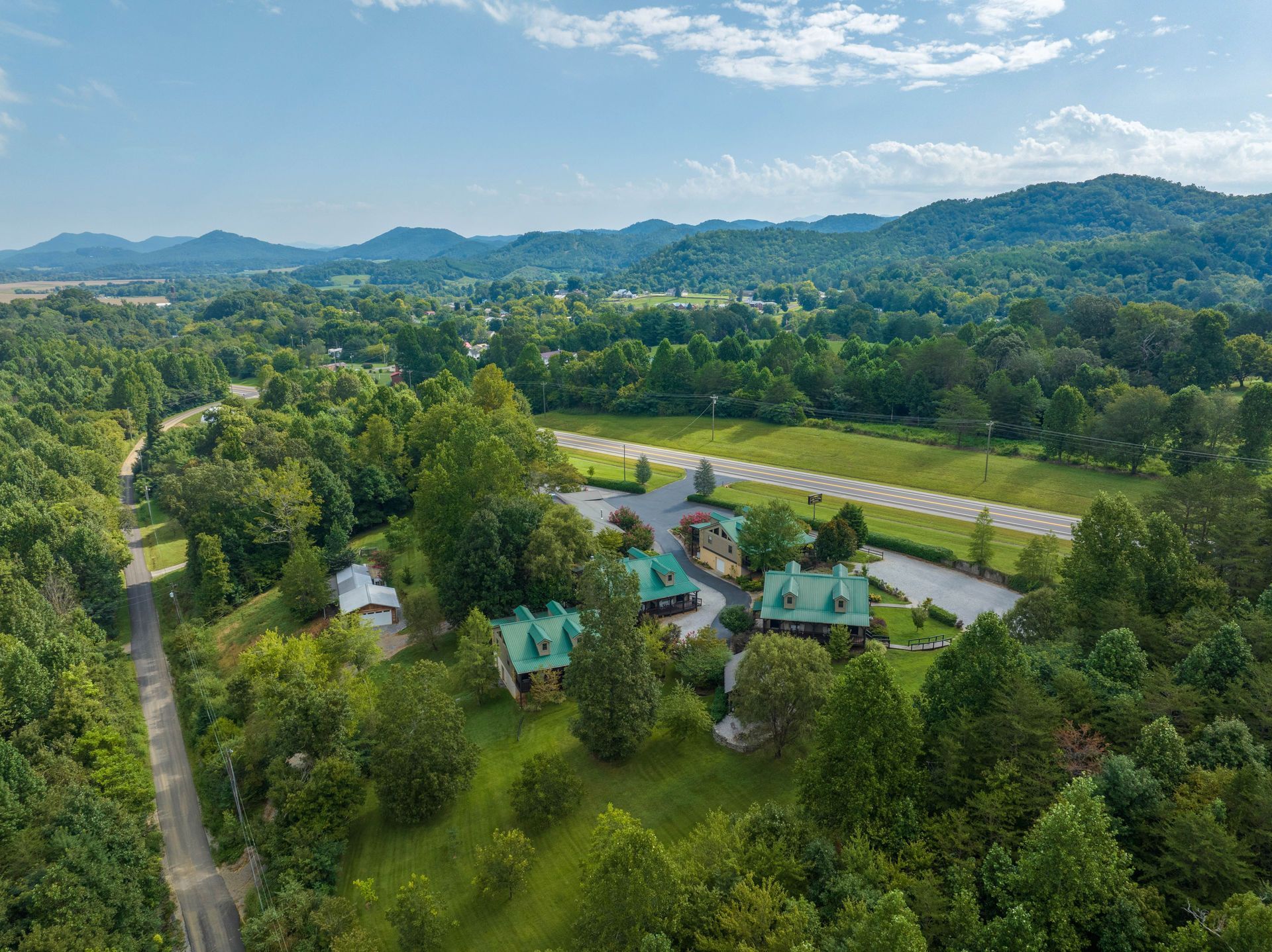 Aerial view of several green-roofed buildings surrounded by trees in a valley with rolling hills in the background.