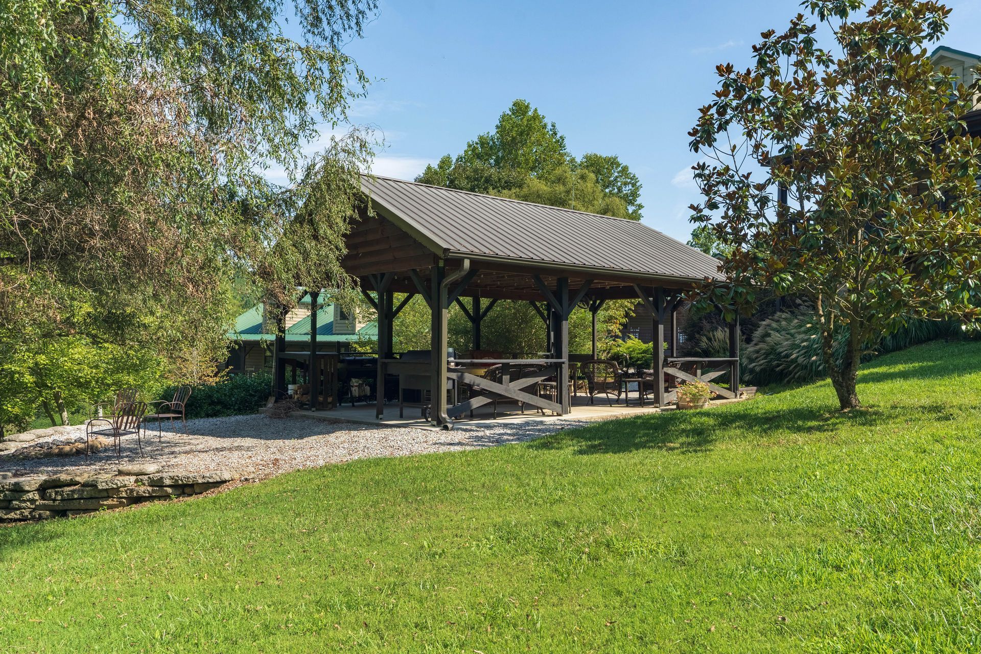 Wooden gazebo with tables and benches, surrounded by green grass, trees, and gravel.