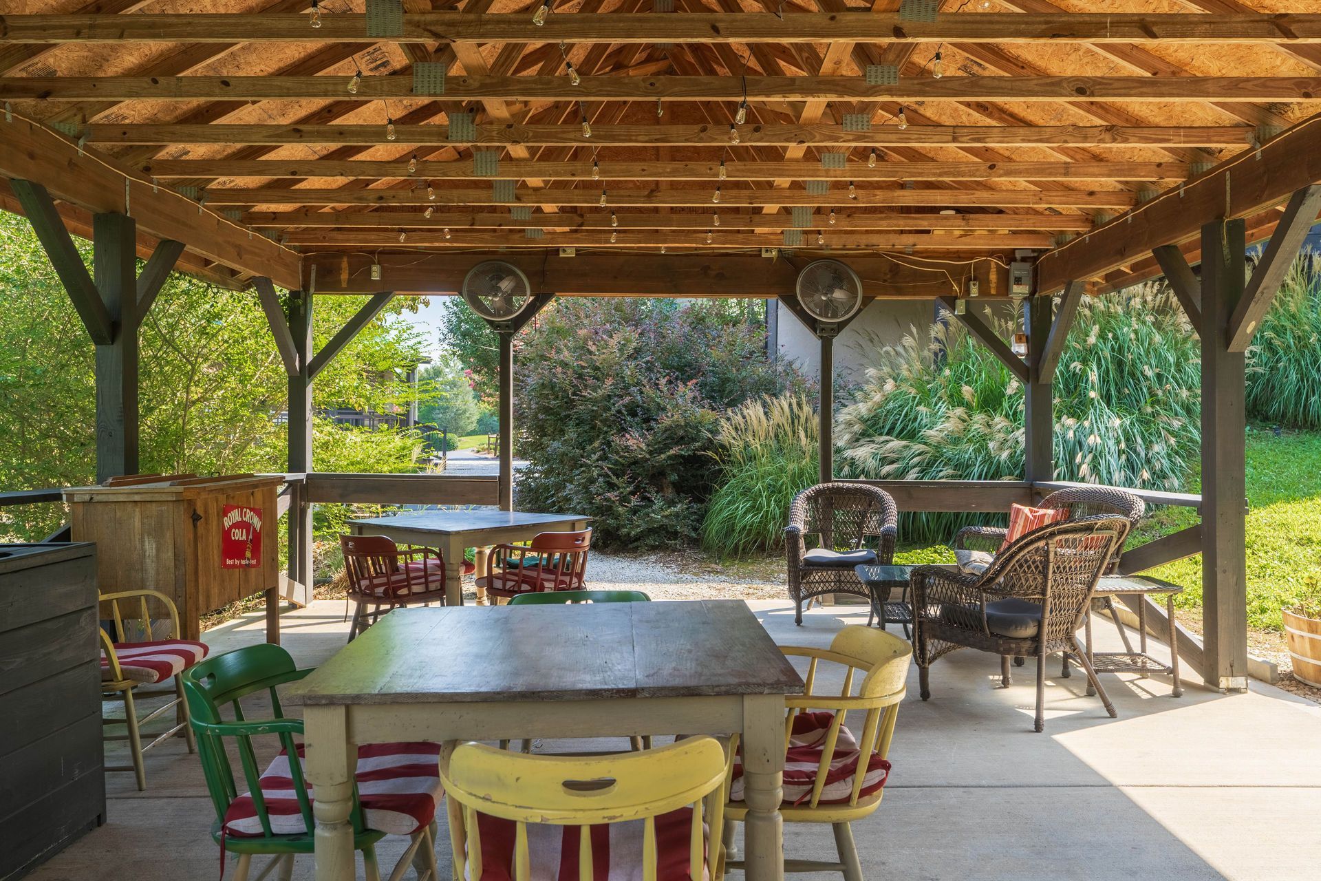 Outdoor seating area under a wooden canopy with tables and chairs, trees in the background.