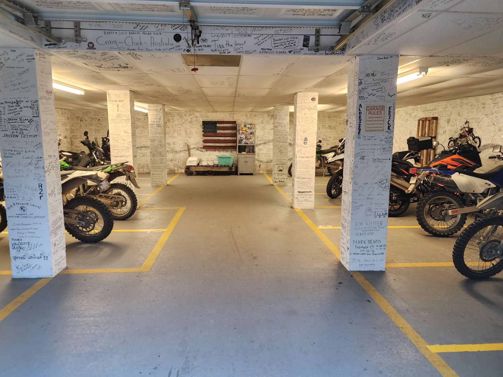 Motorcycles parked in a dim, white-walled garage. American flag and storage against the back wall.