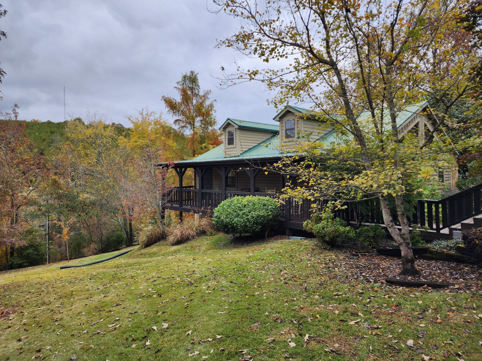 Cabin with green roof, porch, and dormer windows, nestled in fall foliage.