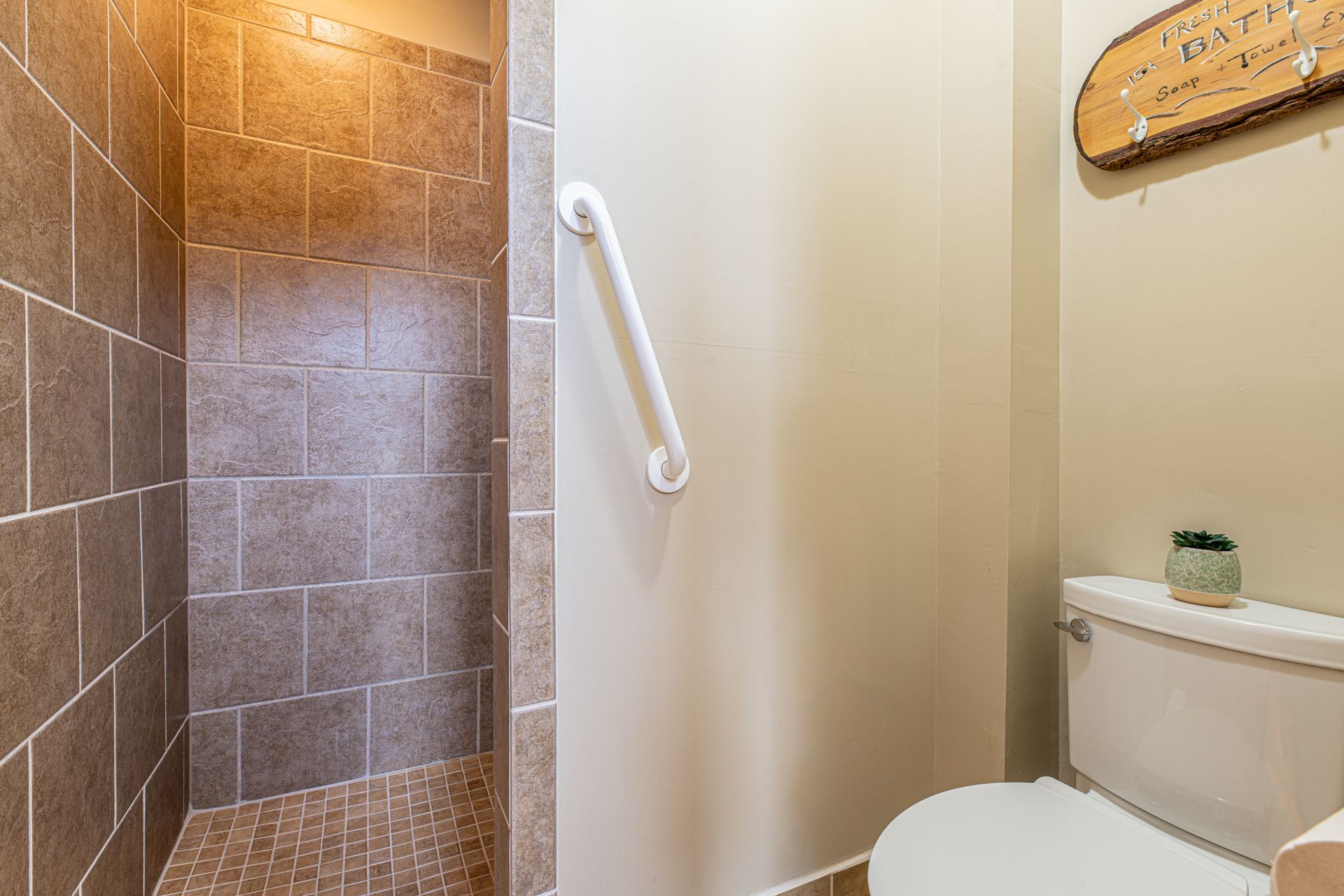 Bathroom with shower, toilet, and grab bar for accessibility. Light brown tile and beige walls.