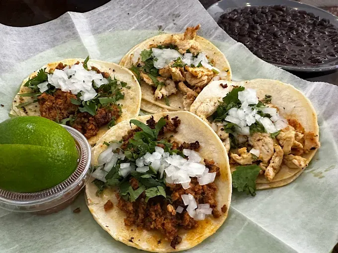 Four tacos with meat, onions, cilantro, and limes, with a bowl of black beans.