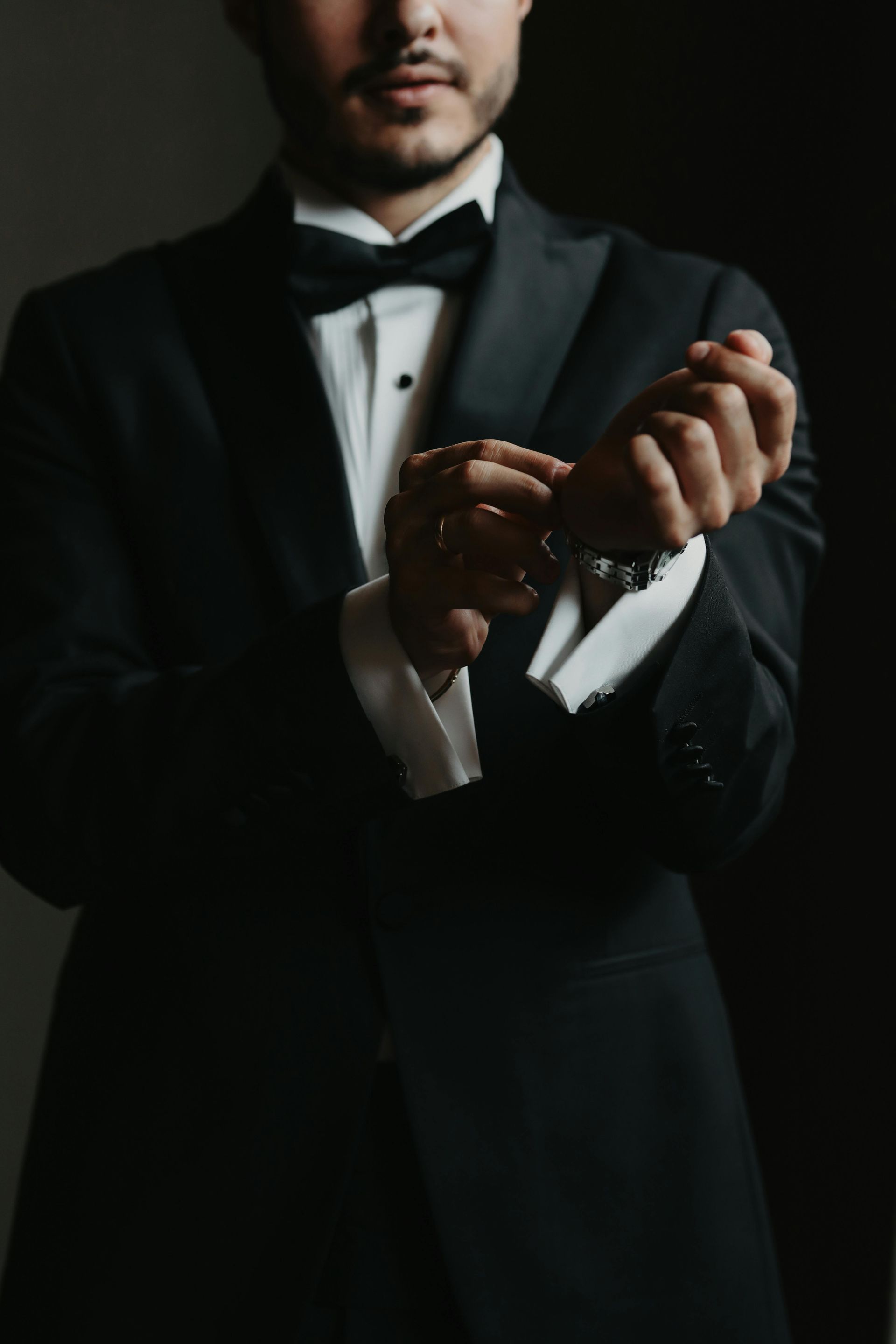 Man in a black tuxedo adjusting his cufflinks against a dark background