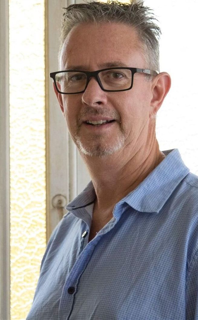A Man Wearing Glasses And A Blue Shirt Is Standing In Front Of A Window — Trinity Family Chiropractic In Cairns, QLD