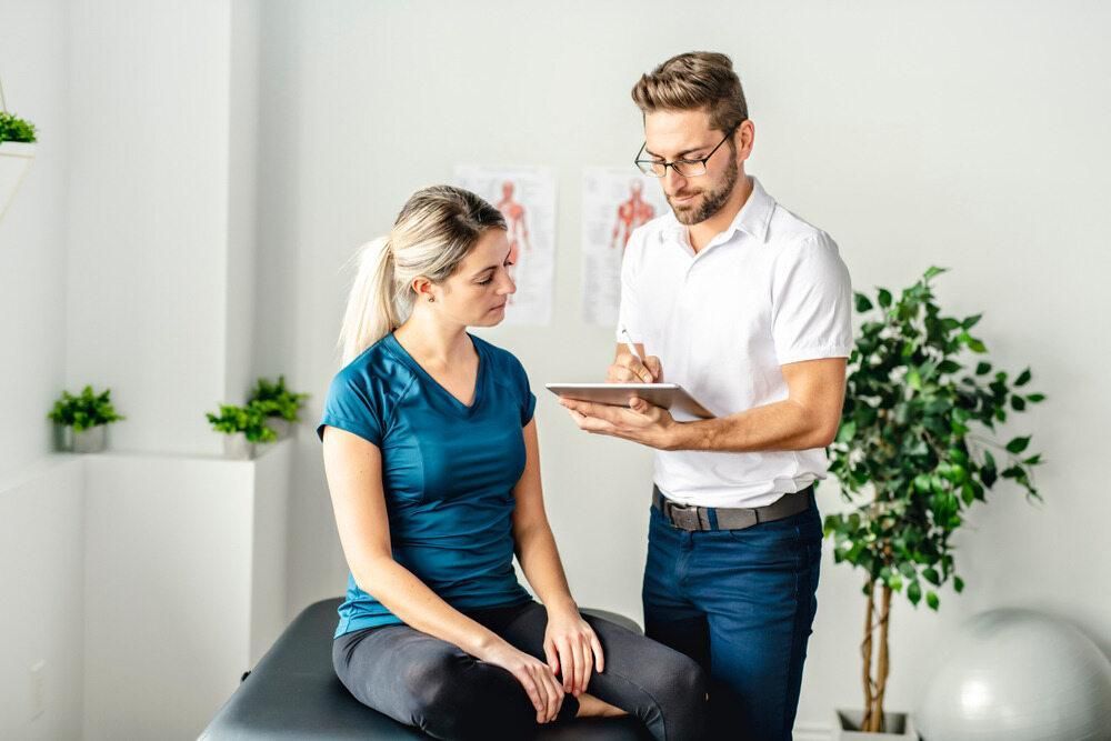 A Man Is Talking To A Woman Who Is Sitting On A Table — Trinity Family Chiropractic In Parramatta Park, QLD