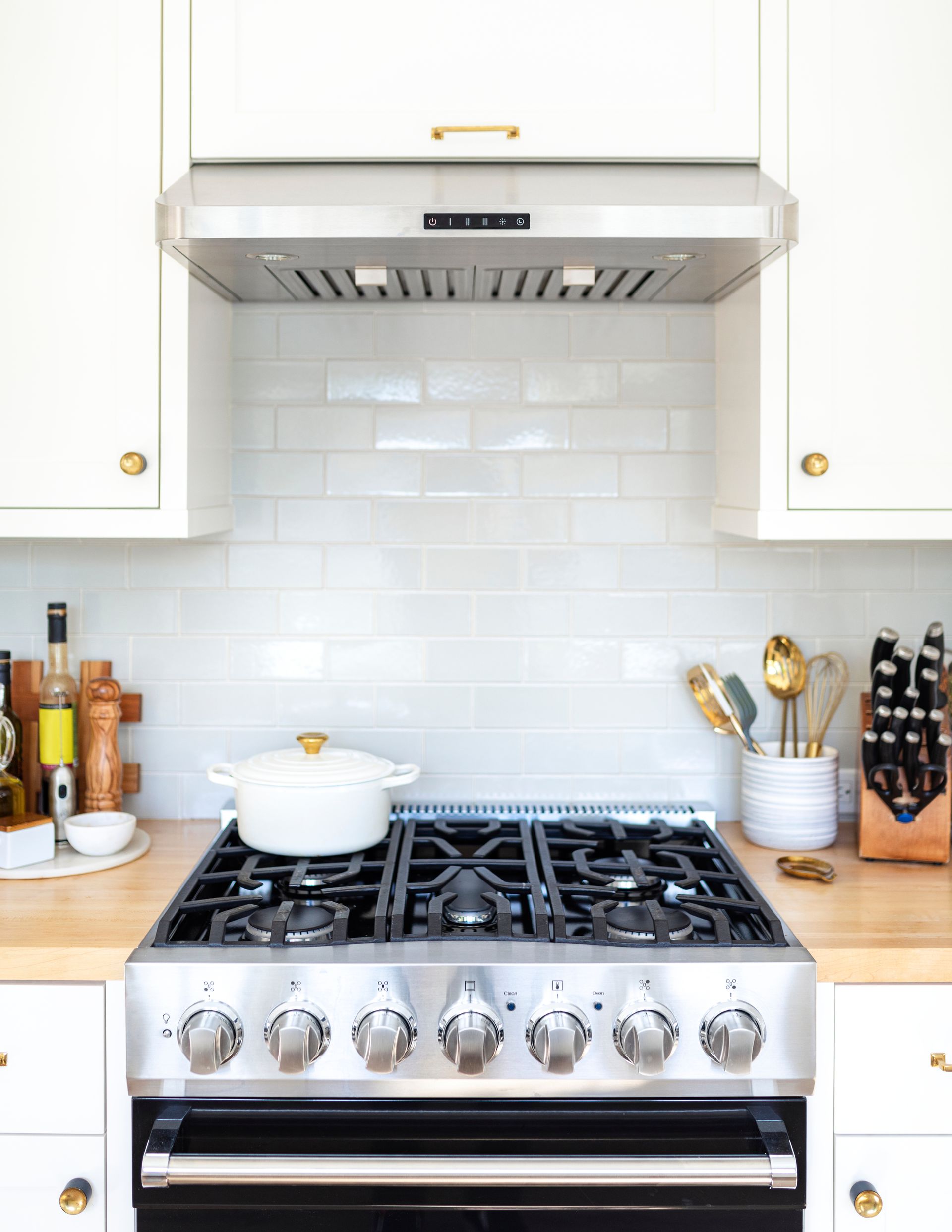 Bright white kitchen with a stainless steel stove, hood, white cabinets, and countertop utensils