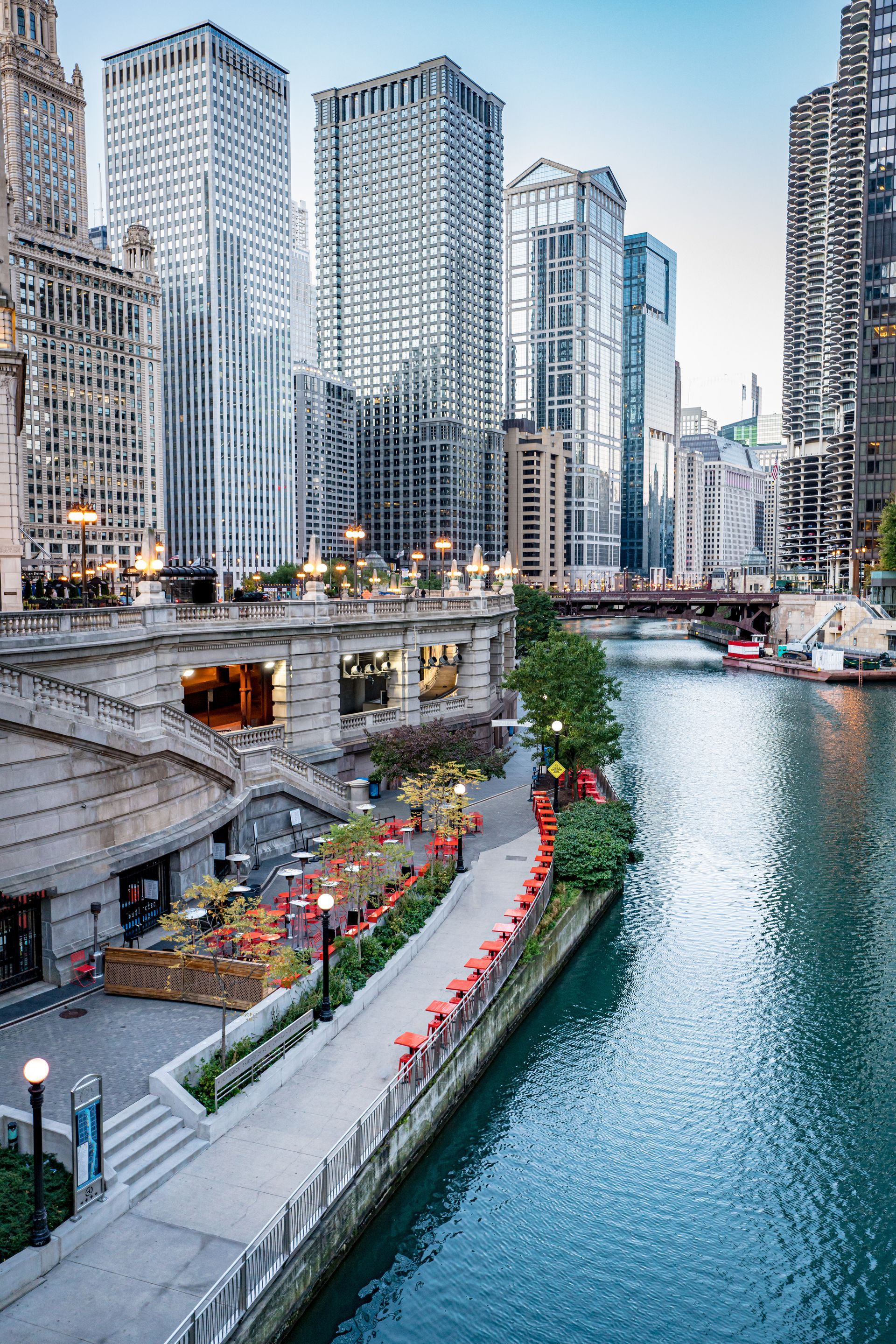 Urban canal lined with flowers and walkway beside tall waterfront skyscrapers at dusk